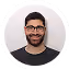 Smiling man with dark hair, beard, and glasses wearing a dark shirt on a gray background.