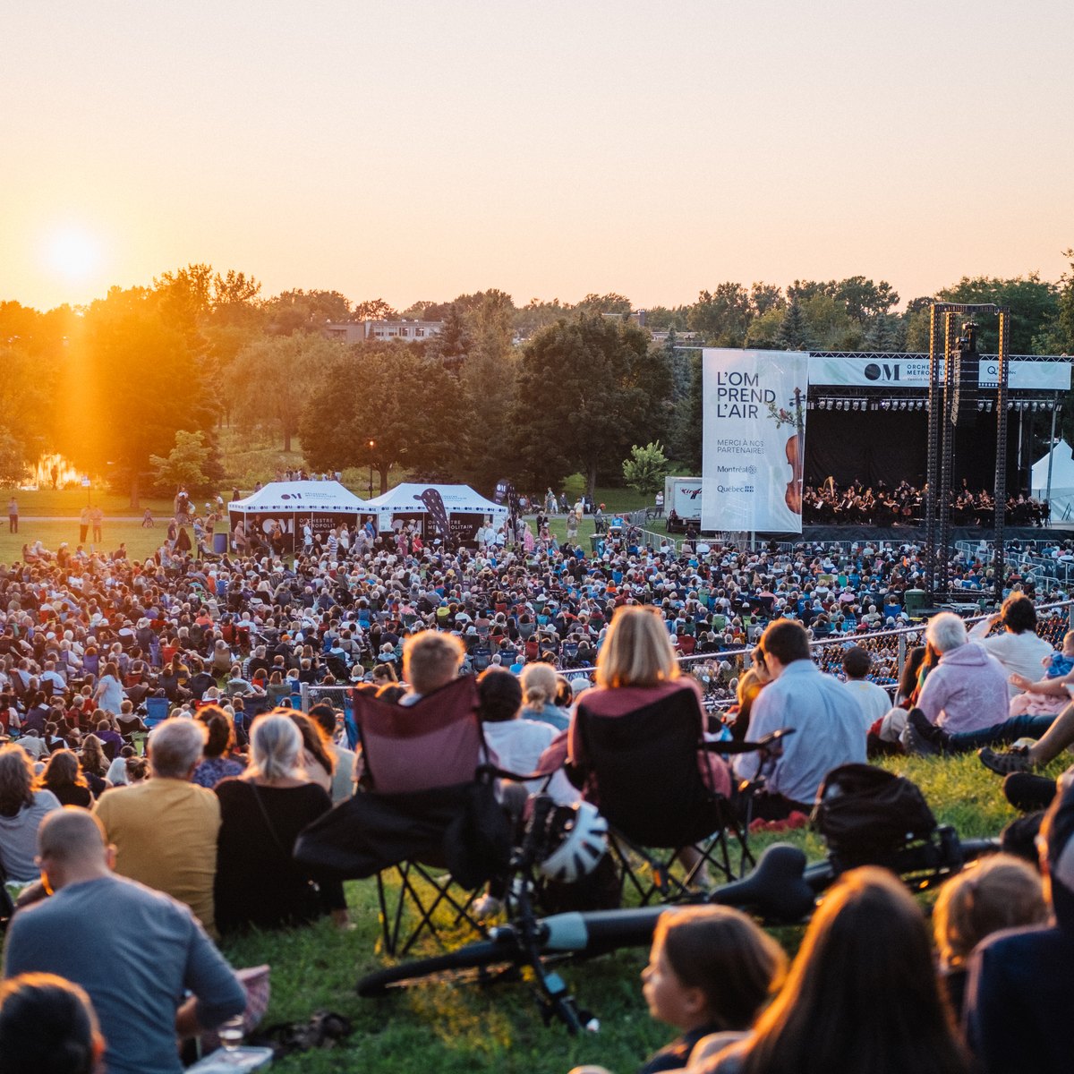 A large crowd seated on the grass at an outdoor concert at sunset