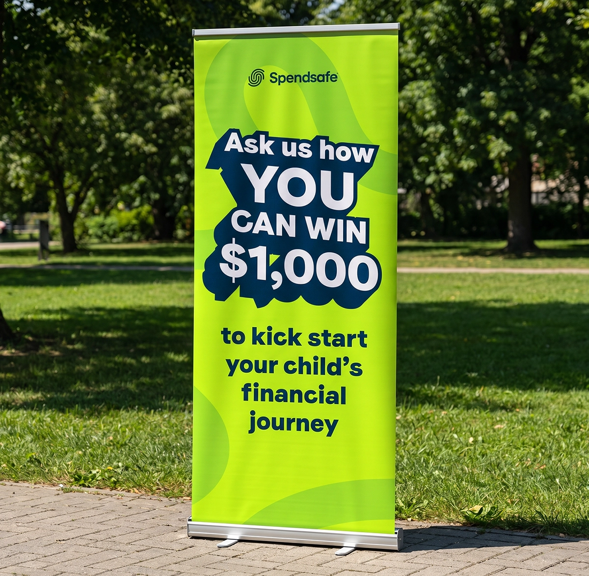 A rollup banner in a park. It reads "Ask us how you can win $1,000 to kick start your child's financial journey."