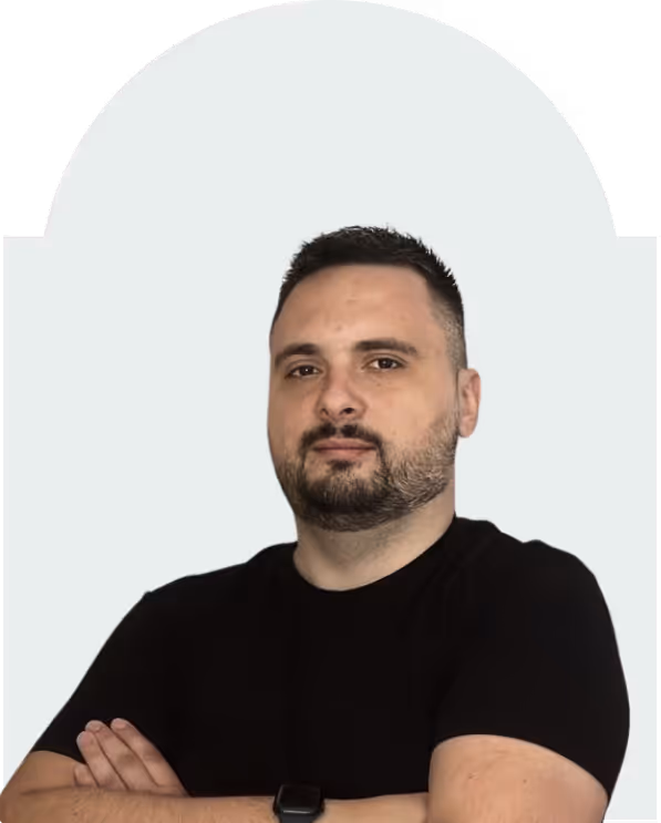Man with short dark hair and beard wearing a black t-shirt and watch, arms crossed, against a light gray background.