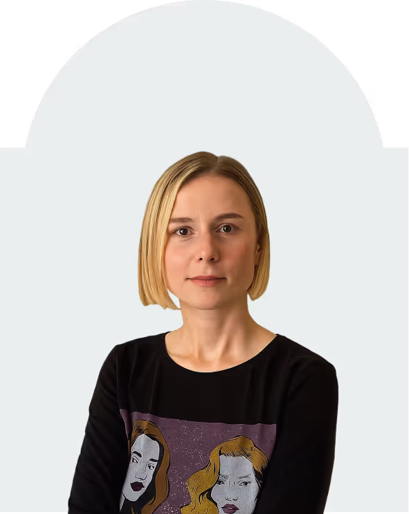 Woman with short blonde hair wearing a black shirt with illustrated faces, standing against a light gray background.