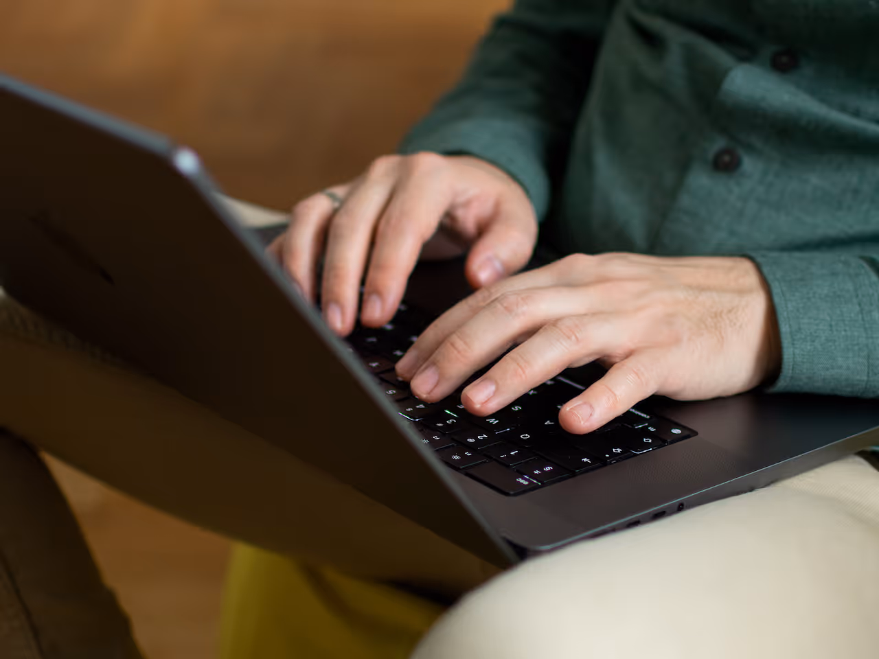 Person wearing a green shirt typing on a black laptop keyboard with beige pants visible.