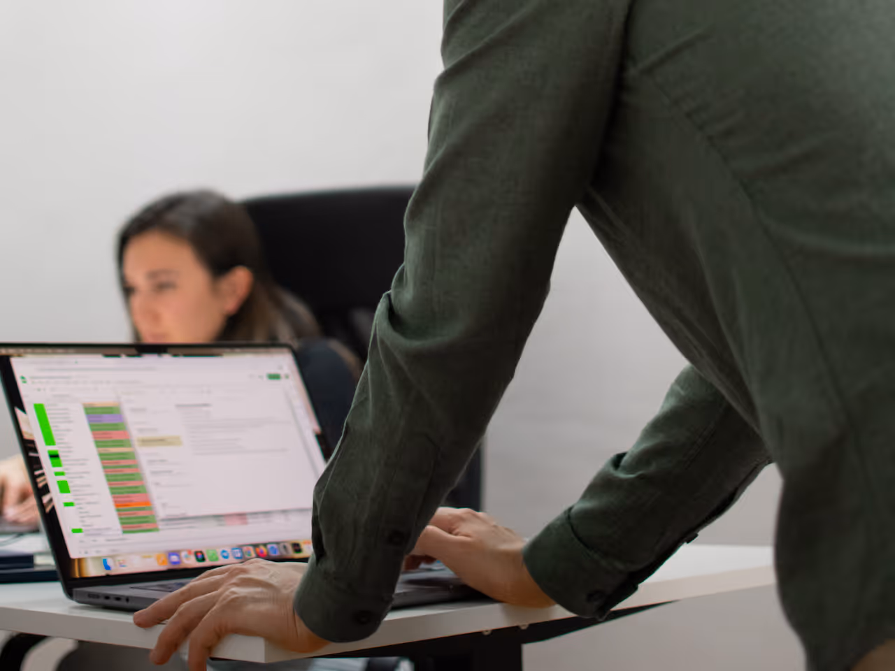 Person in green shirt typing on a laptop displaying a color-coded spreadsheet while another person sits blurred in the background.