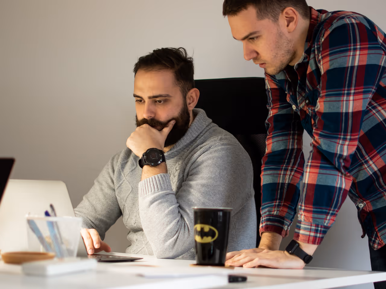 Two men focused on a laptop screen at a white desk, one seated in a gray sweater and the other standing in a red and blue plaid shirt.