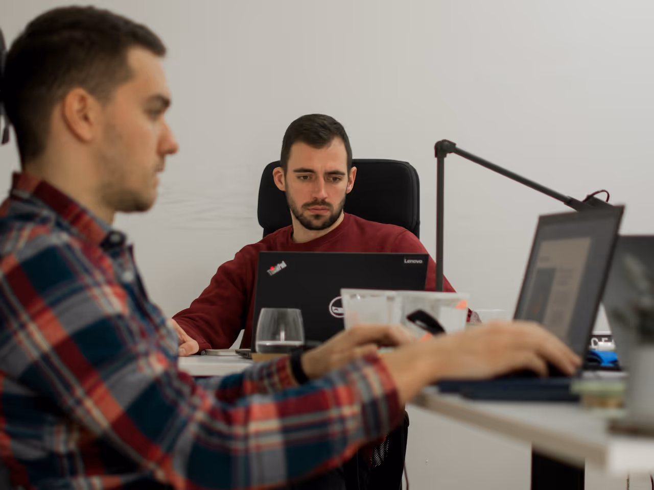 Two men working on laptops at a shared office desk with a white wall background.
