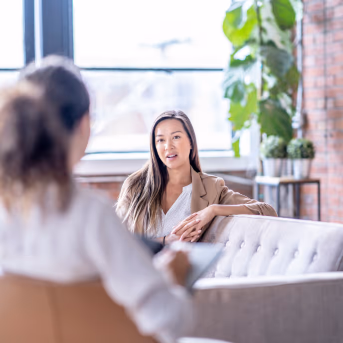 A woman sits on a sofa in a bright, modern office, engaged in a mental health therapy session. The therapist takes notes as they talk in a calm setting with natural light and indoor plants.    