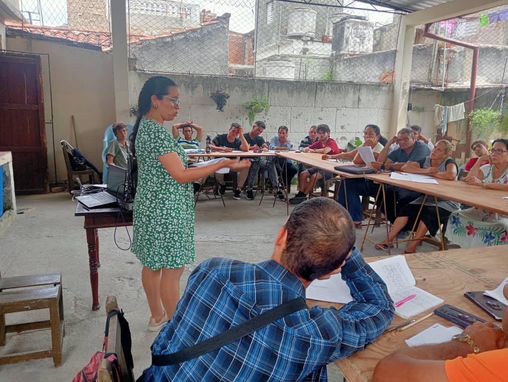 A woman leads a business training class for Cuban adults seated at long tables in an outdoor courtyard