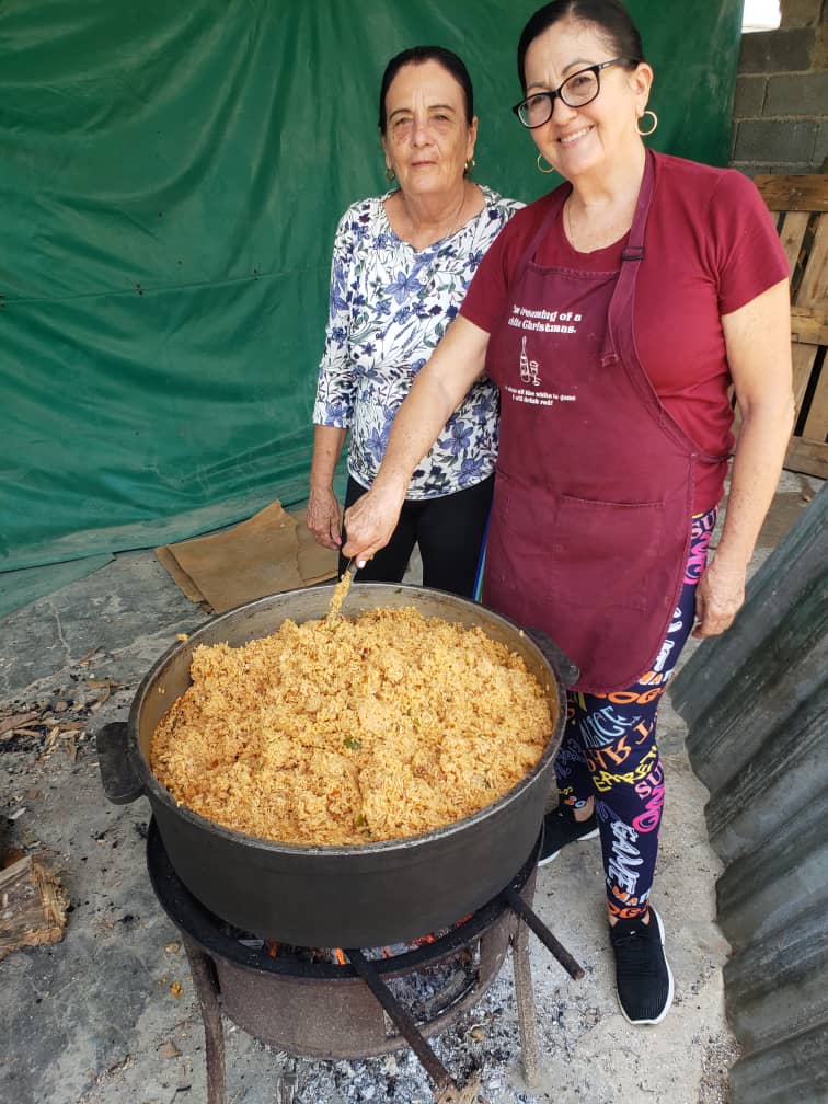Two women smile beside a large pot of rice cooking over an open fire