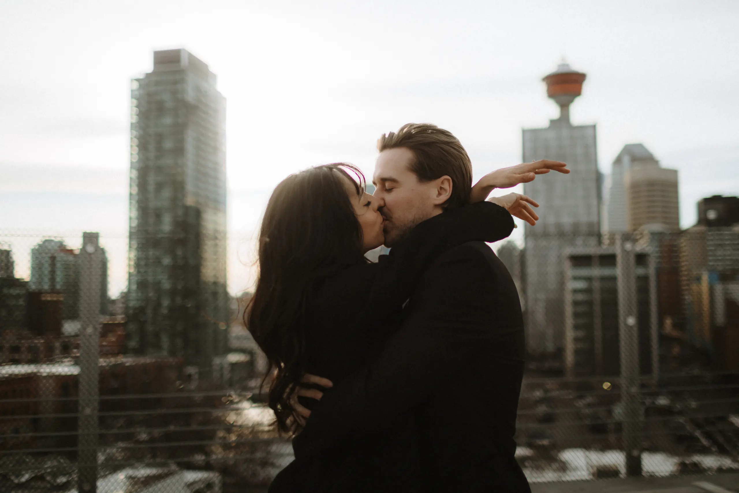 Engaged couple kissing on top of a parkade surrounded by skyscrapers downtown Calgary