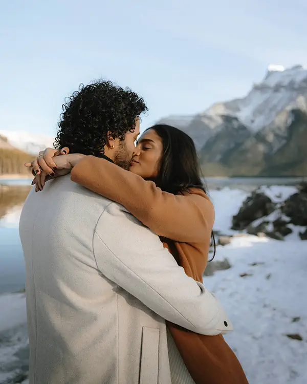 Couple embraces beside Lake Minnewanka during proposal photoshoot with snow-capped Canadian Rockies backdrop