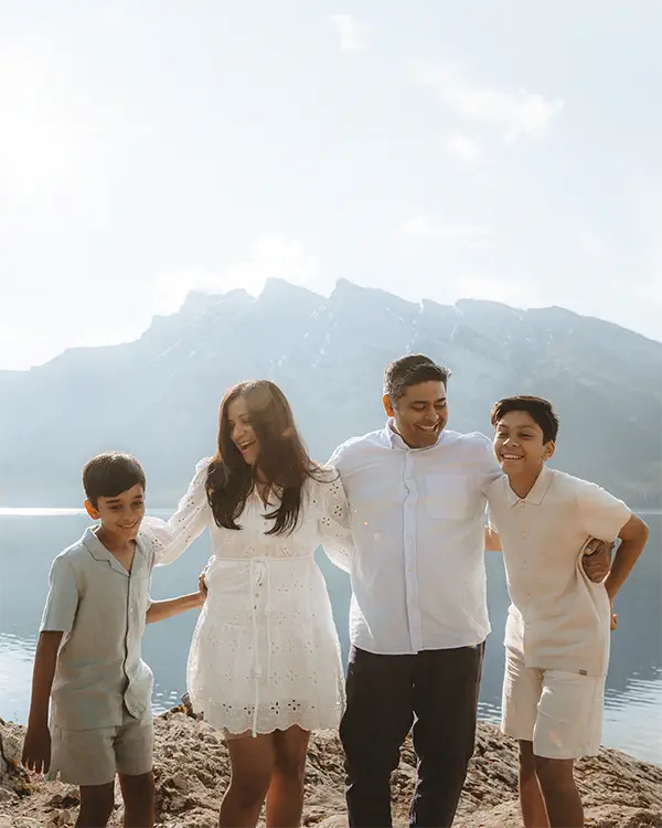 Happy family of four poses together on rocky shoreline of Lake Minnewanka with massive mountain backdrop in Banff