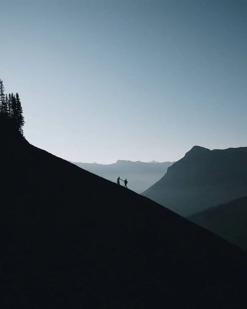 Two adventurers hold hands as silhouettes against dramatic mountain slope in Canadian Rockies wilderness