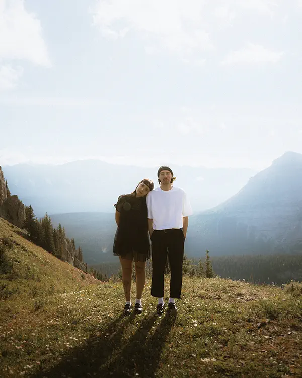 Couple stands, leaning on each other on Banff mountain summit with layered ridges stretching into distance