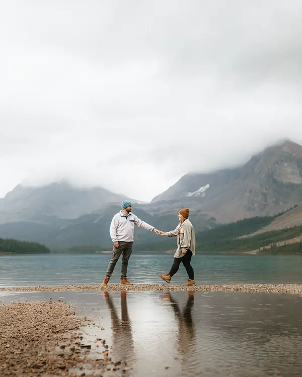 Couple walks hand in hand along Bow Lake shore with snow-capped peaks reflected in water