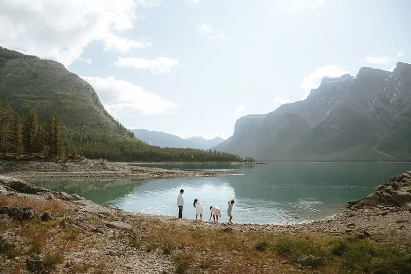 Family explores pristine mountain shoreline of Lake Minnewanka surrounded by towering peaks and evergreen forest