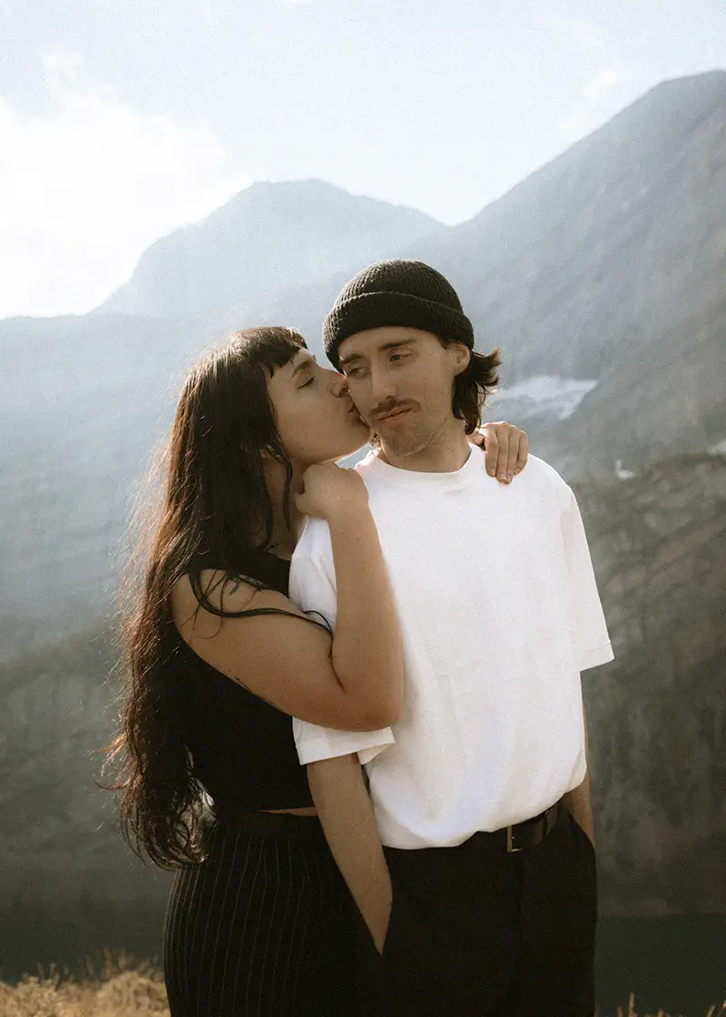 Woman kisses partner's cheek in tender moment with misty Kananaskis mountain valley behind them