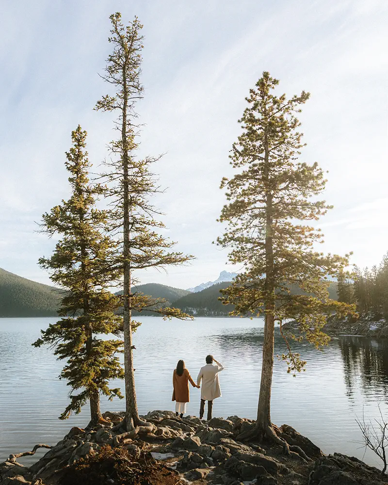 Couple stands beneath tall pine trees beside calm Lake Minnewanka during golden afternoon photoshoot