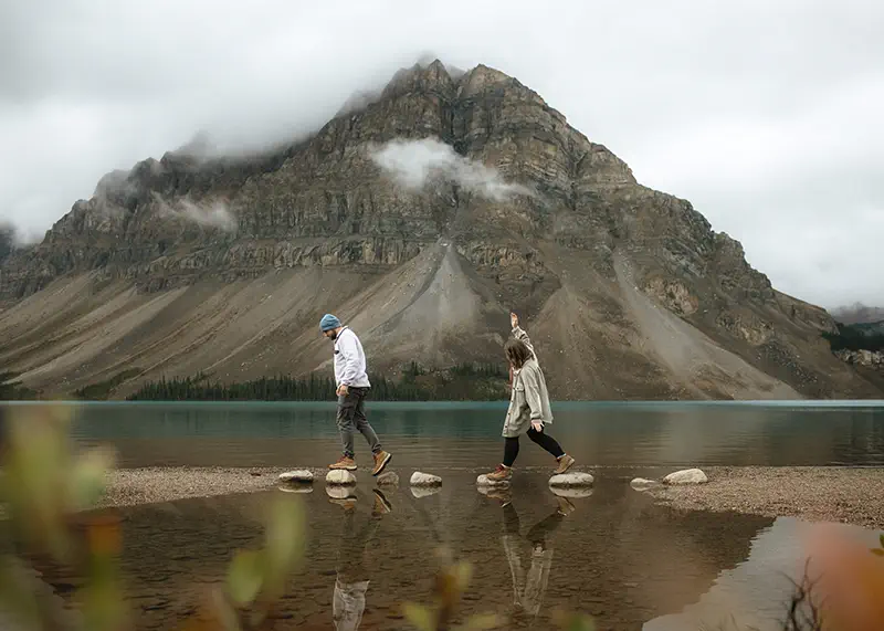 Playful couple crosses mountain lake on stepping stones with dramatic cloudy peak backdrop