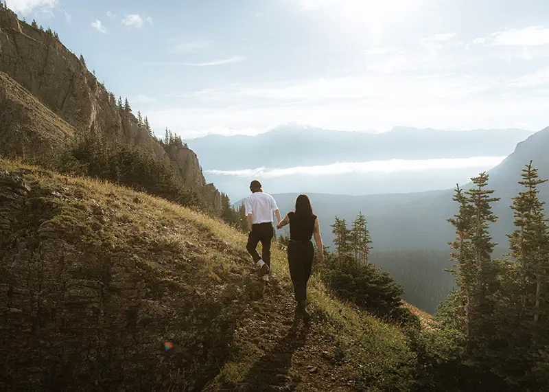 Couple hikes up mountain trail hand in hand with Canadian Rockies valley views stretching behind them