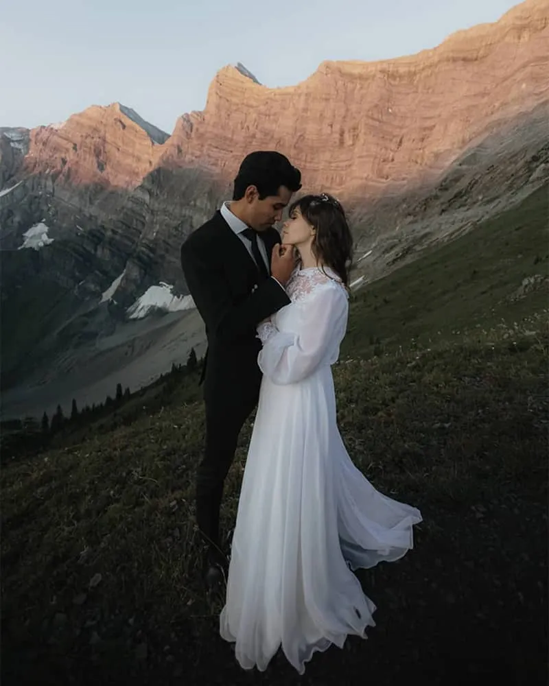 Bride and groom embrace in alpine valley with dramatic rocky peaks at sunrise in Kananaskis, Alberta