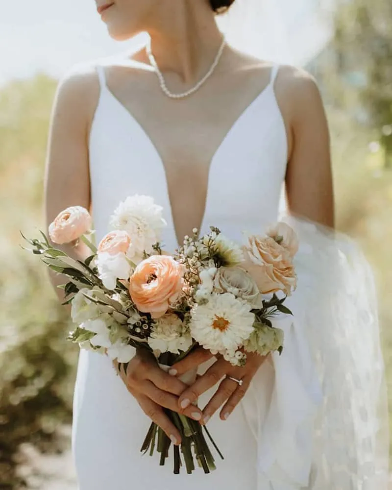 Bride in gown holds soft peach and white bridal bouquet for intimate wedding ceremony