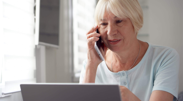 An elderly lady holding her phone to her ear sitting in front of a laptop.