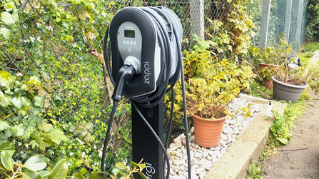 Electric vehicle charging station with coil of cable mounted on a black stand next to a garden with potted plants and greenery.