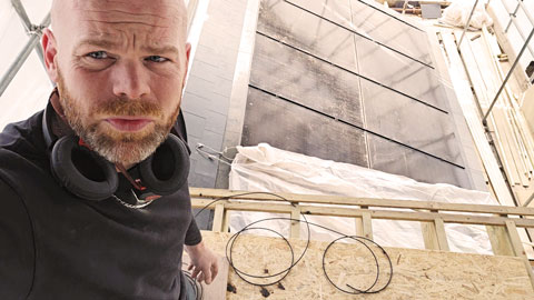 Man with headphones around his neck working on a wooden frame near black solar panels on a roof.