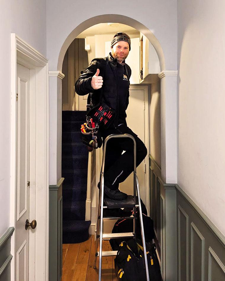 Man wearing a beanie and work clothes standing on a ladder in a hallway giving a thumbs-up.