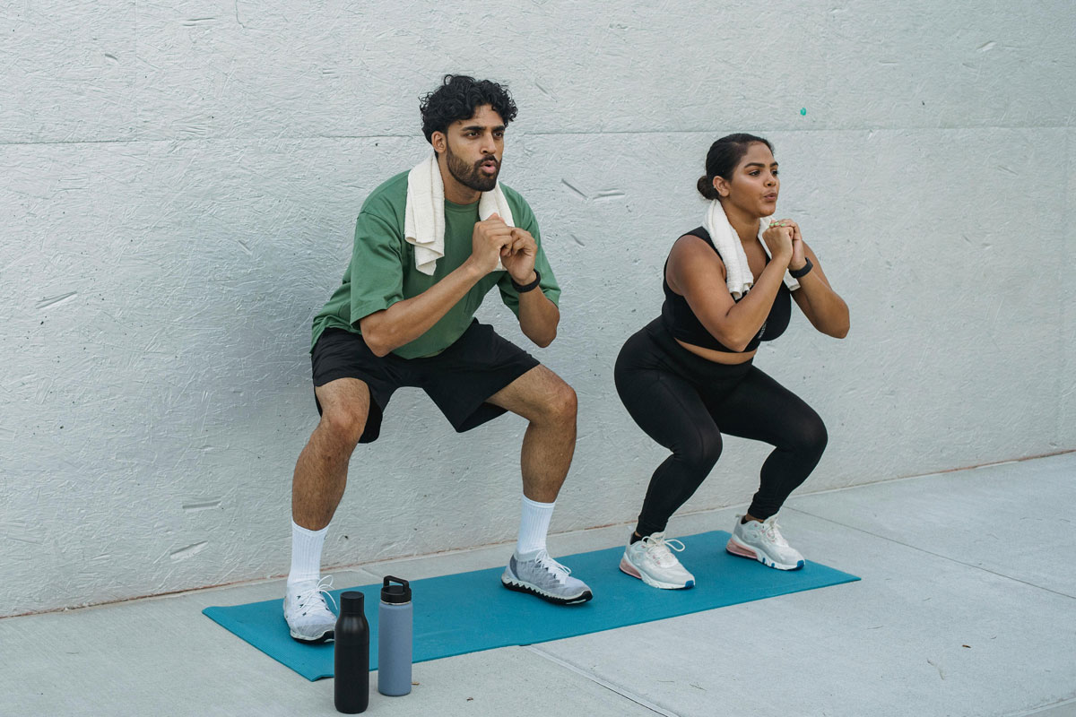 A man and a woman perform synchronized squats outdoors on a blue yoga mat, both wearing athletic wear and white towels around their necks, representing a fresh spring workout.