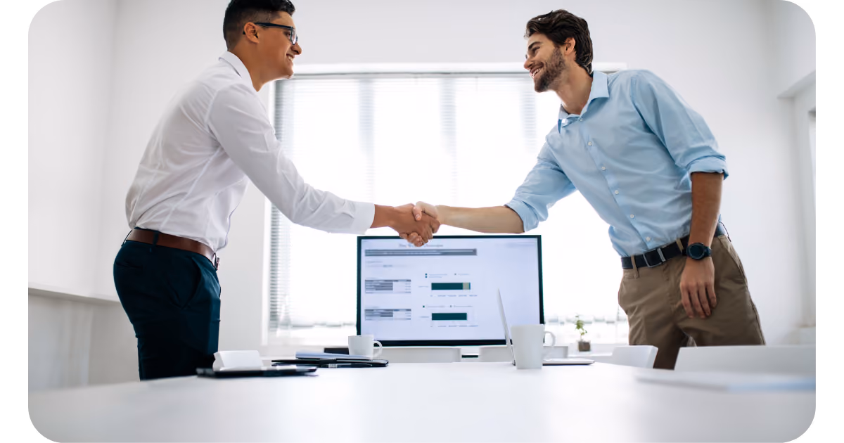 Two people shaking hands across a white desk with a computer monitor displaying data