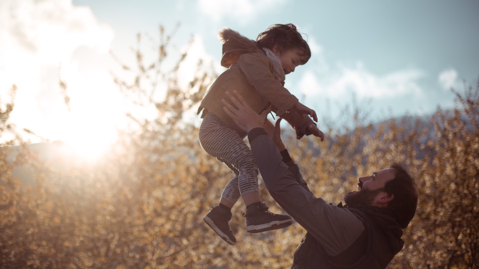 Father holding his child up. They are in a field. The sun is setting behind them