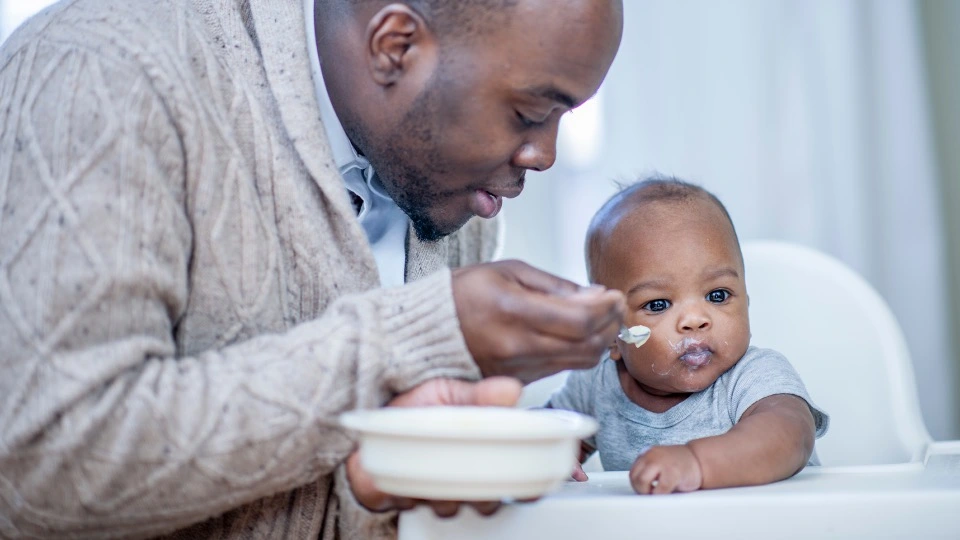 Father feeding his child who is sitting in a high chair.