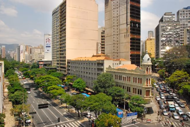 City street with tall buildings, trees lining roads, and cars in traffic under a cloudy sky.