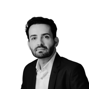 Man with dark hair and beard wearing a white shirt and black suit jacket on a white background.