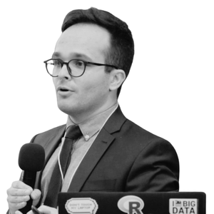Man in suit and glasses speaking into microphone behind a laptop with data and R stickers.