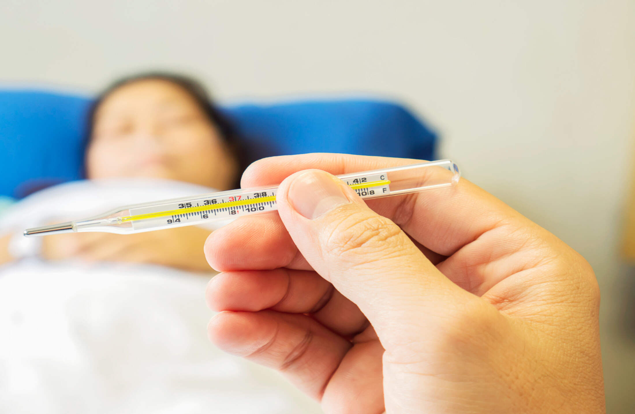 Hand holding a glass thermometer showing a fever, with a blurred person lying down in the background.