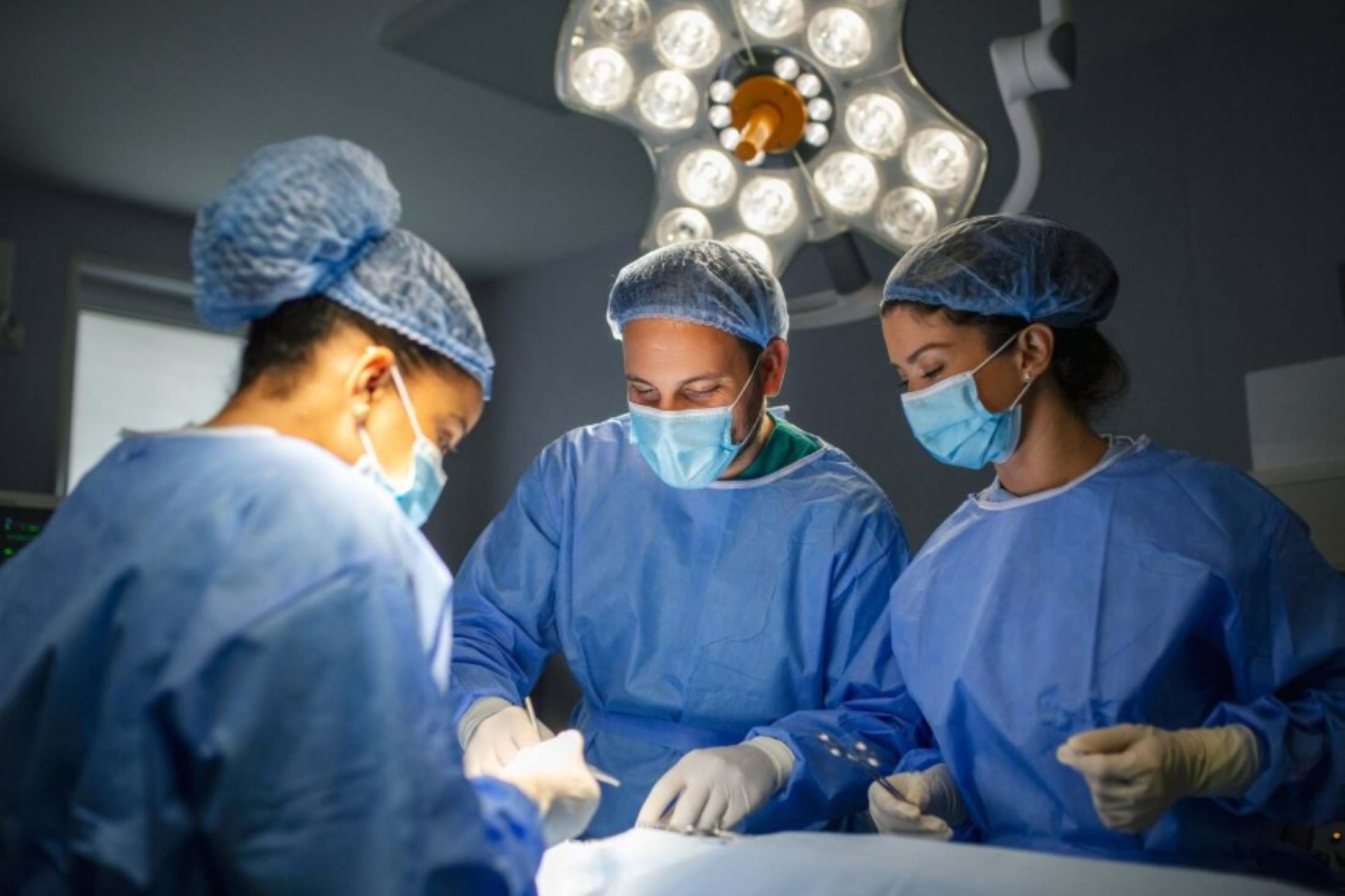 Three surgeons in blue scrubs and masks performing surgery under bright operating room lights.