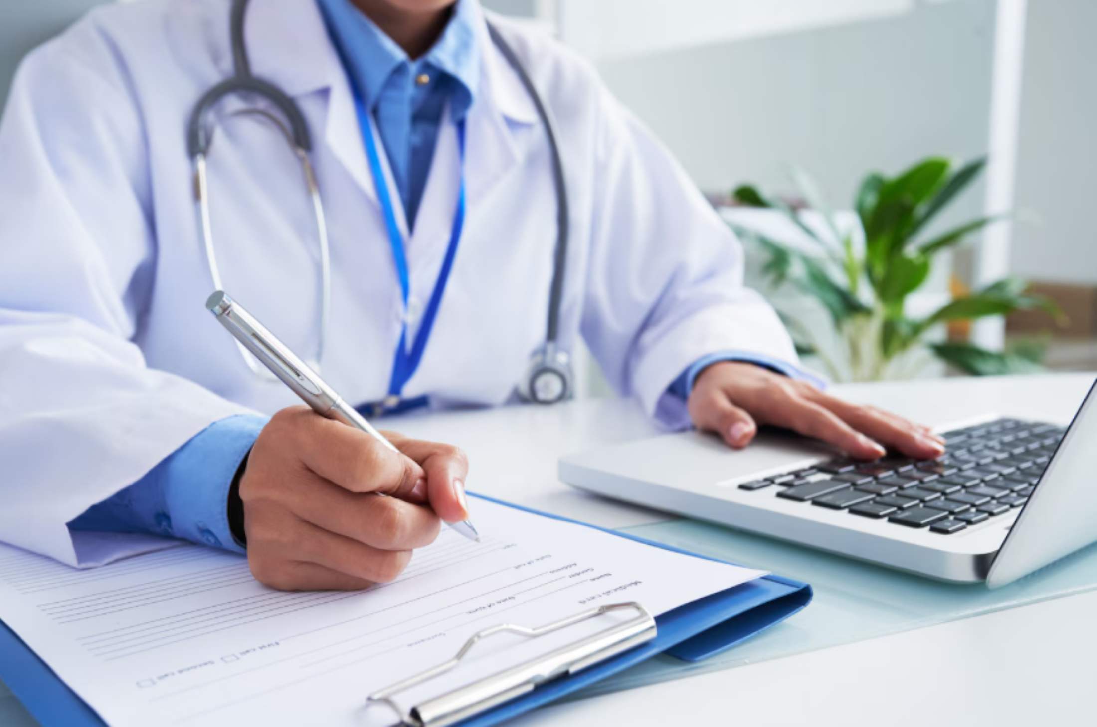 Doctor in white coat writing on clipboard with one hand and typing on laptop with the other.
