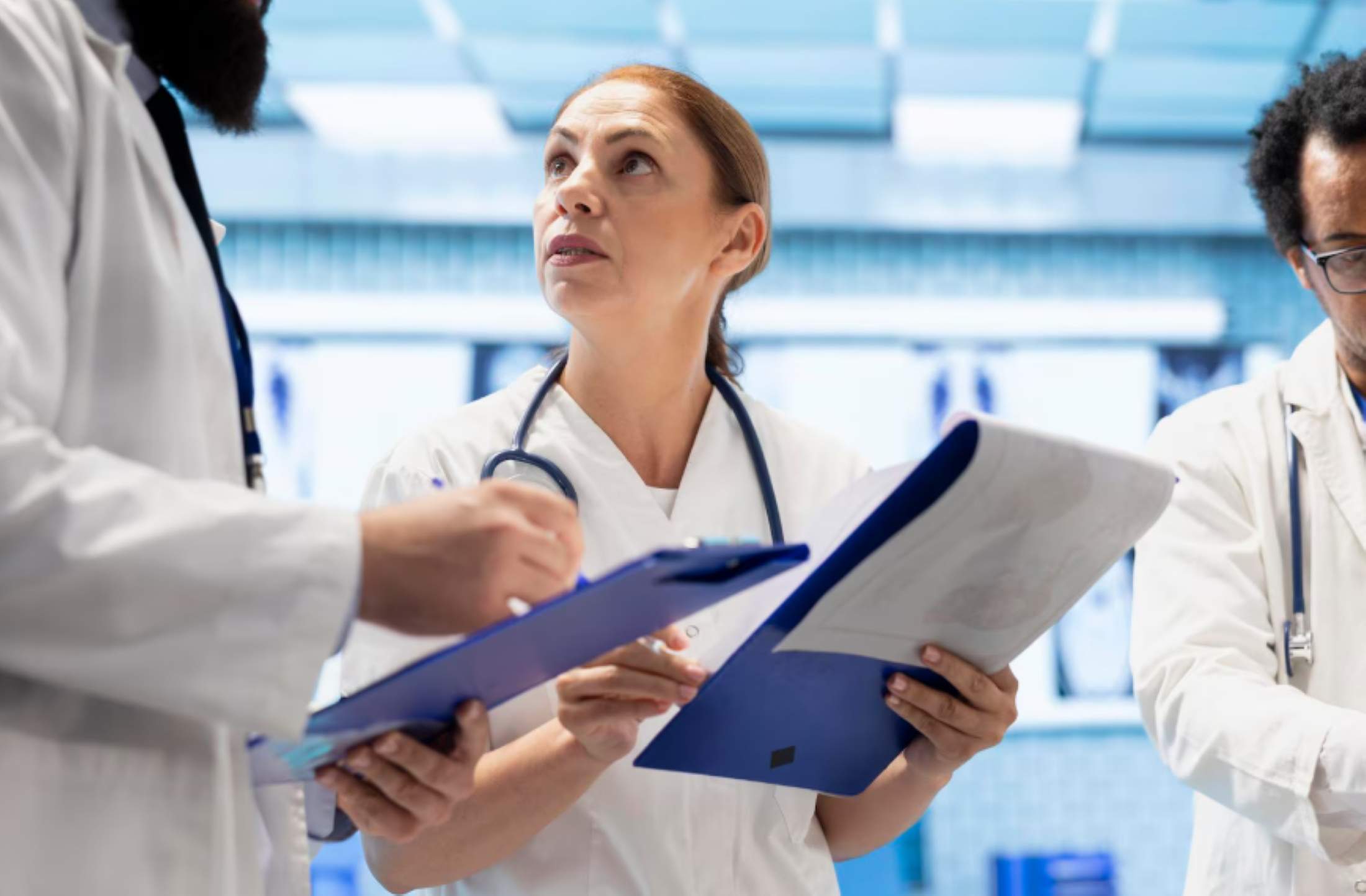 Three doctors in white coats with stethoscopes discussing notes on clipboards in a medical setting.
