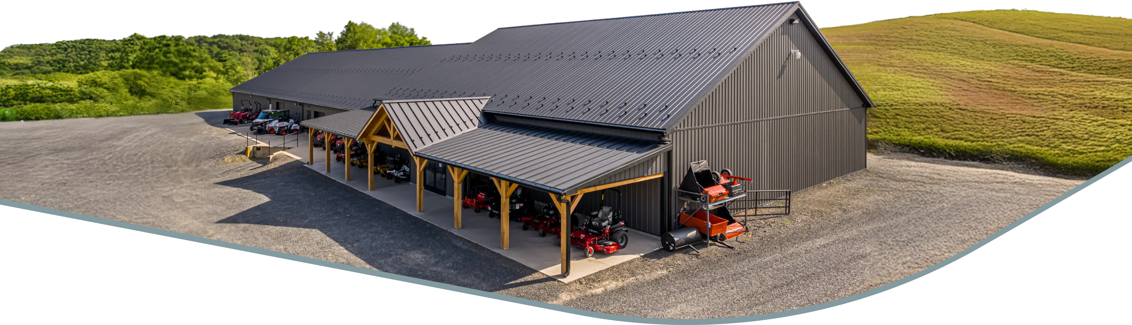 A large metal building with a covered area displaying various farm equipment and machinery on a gravel lot, set against a backdrop of trees and an open field.