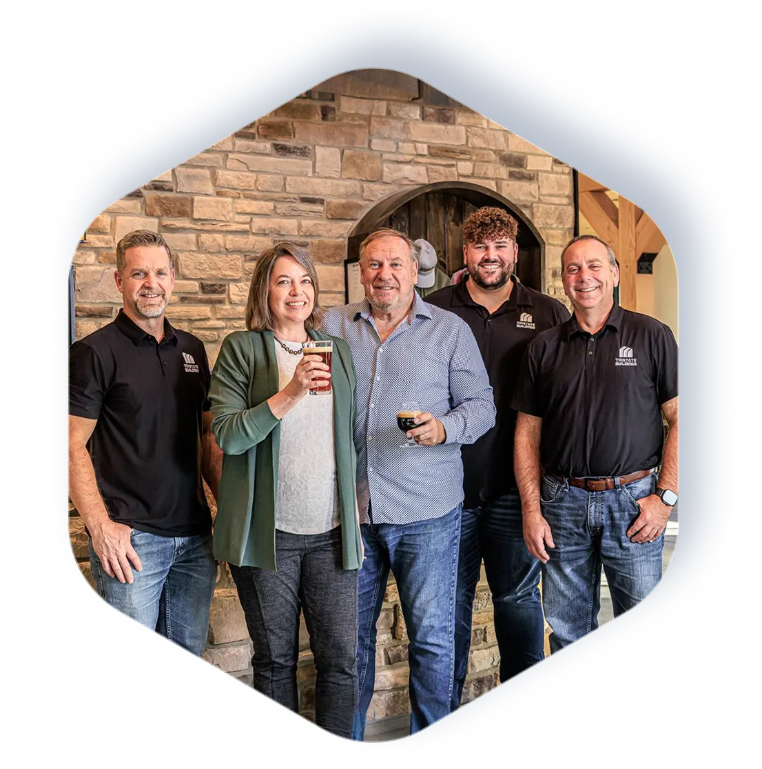 Tristate team standing indoors against a stone wall, three men wear TriState Builders shirts, two hold drinks.