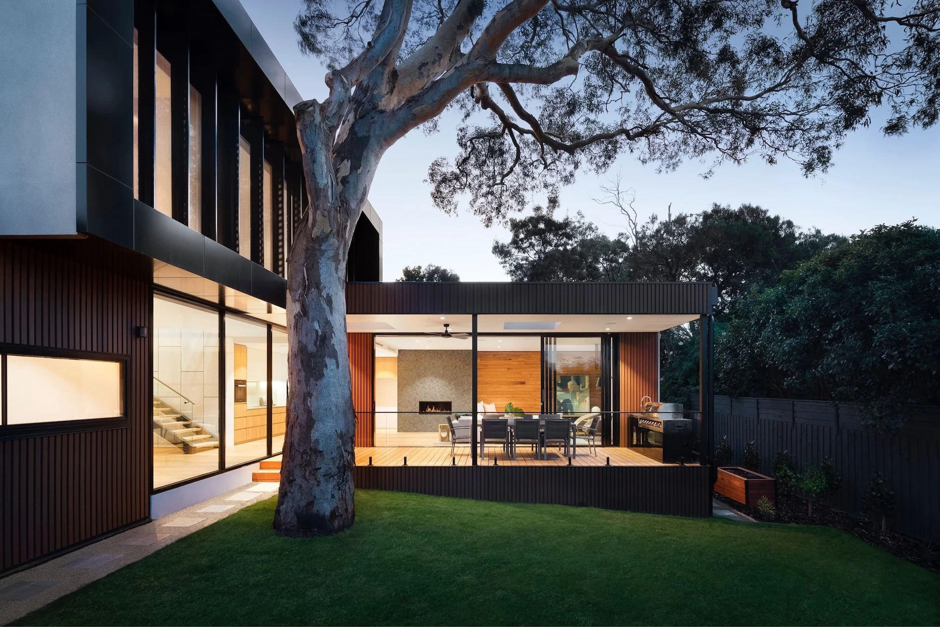 Modernes Haus mit großen Glasfronten und Holzverkleidung, im Vordergrund ein großer Baum und gepflegter Rasen, dahinter eine überdachte Terrasse mit Sitzbereich.
