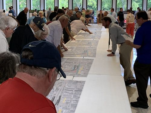 A group of people in the cafeteria of St. Thomas More High School on August 2, 2023 for the public involvement meeting. Individuals are viewing proposed design concepts displayed on roll plots across a long table.