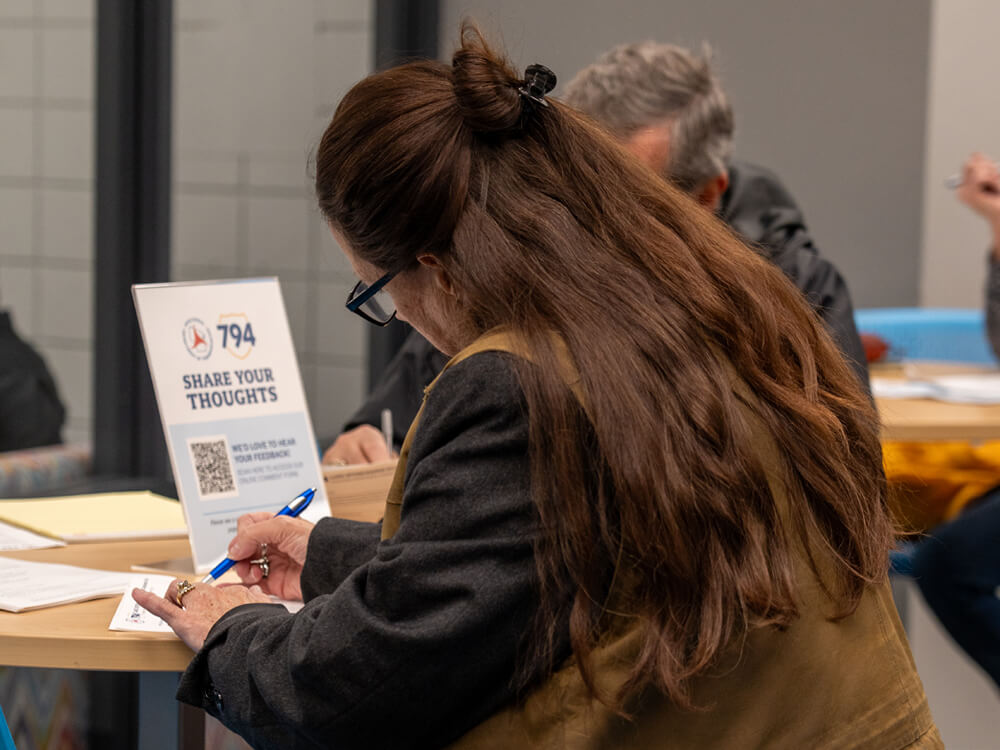 An attendee writes feedback on a form next to a sign reading ‘Share Your Thoughts,’ which includes a QR code.