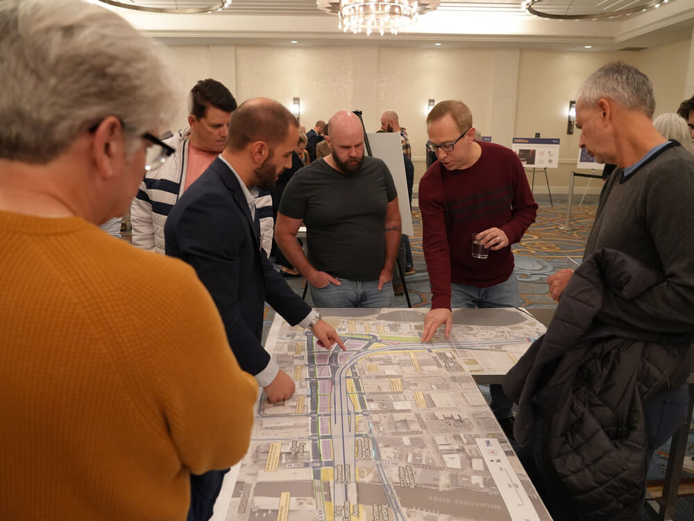 A small group of attendees gathers around a large, printed roadway map laid out on a table. One participant points to a section of the map while others review project details and discuss proposed corridor changes during the public information meeting.