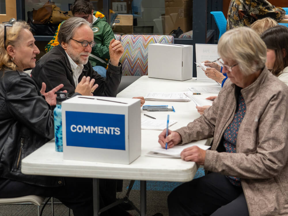 Visitors sit at a table filling out written feedback forms during the public information meeting. A box labeled ‘Comments’ is placed at the center of the table, and attendees write while discussing the project