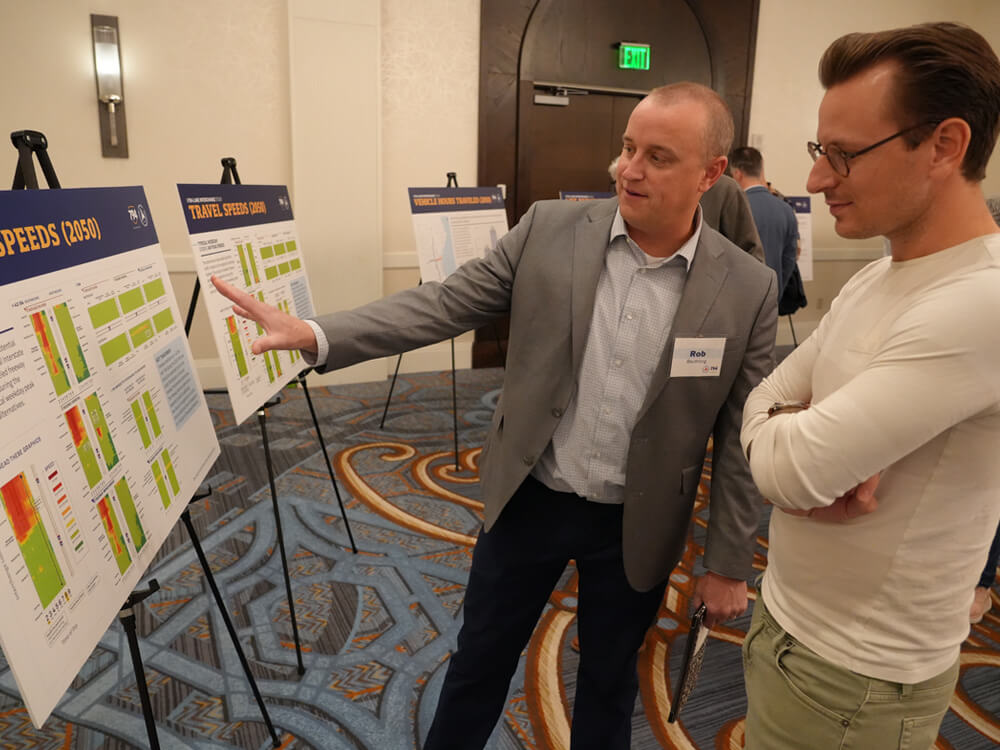 A project representative gestures toward a display board showing traffic speed charts and maps while explaining findings to an attendee. Multiple easels display project data as part of the public information meeting.