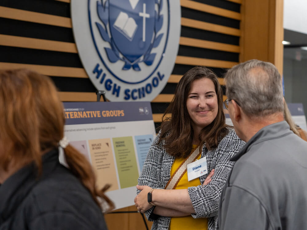 A woman smiles while speaking with an attendee during the public information meeting at St. Thomas More High School. A large school emblem hangs on the wall behind her, and a display board outlining project alternatives is set up nearby.
