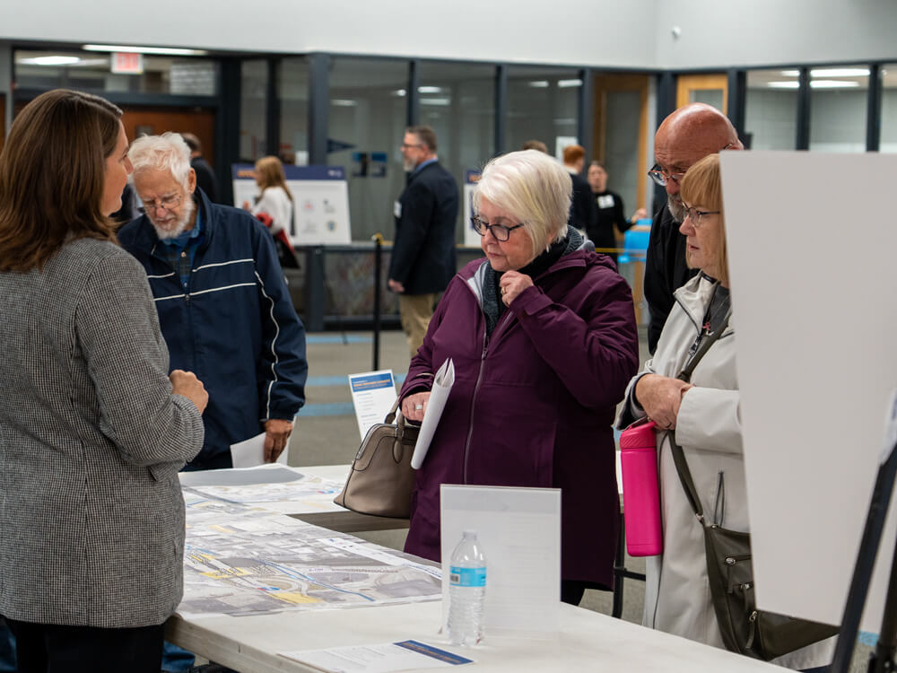Attendees stand at a table covered with detailed roadway maps and printed materials. A project representative answers questions while participants review drawings and hold reference packets during the public information meeting.
